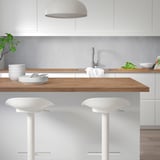 Modern kitchen with white counter, wooden decor, minimalist stools, sleek sink, and pendant light.