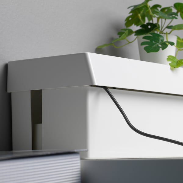 White box, likely the SÄTTING, concealing cables. Lid designed for easy cable access. Plant nearby, cable visible.