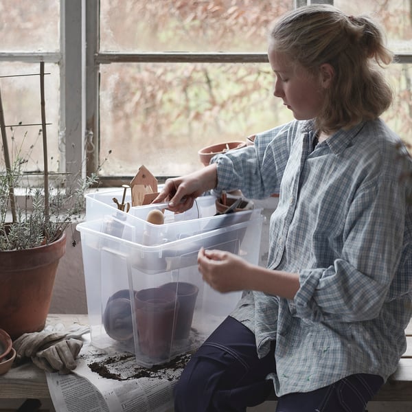 Woman sits by window, handling crafts in clear plastic SAMLA box. She wears a checkered shirt, focusing on miniatures. Potted plant and gloves nearby.