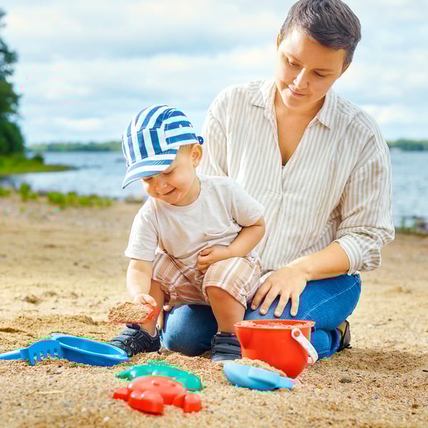 A woman and child play with sand toys on the beach. They use a bucket, rake, and spade, enhancing motor skills and creativity.
