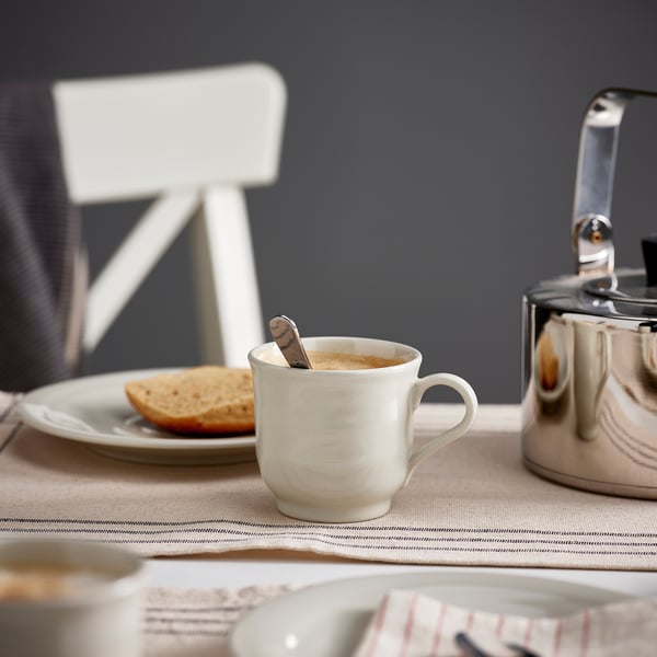 A white ceramic mug filled with coffee sits on a beige tablecloth near a metallic kettle and a small plate with toast.