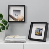 Two black frames on shelf: one with seashell, feather, photo; other with drawing. Magazines and plant nearby.