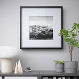 A framed black-and-white rocks sits on a cabinet, surrounded by a lamp, plants, and decorative stones. The frame displays the photo prominently.