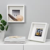 White shelves with framed items: a seashell photo and a colorful building print, alongside potted plant and starfish.