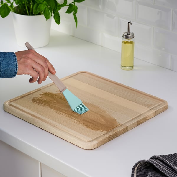 Hand brushes oil on wooden cutting board, with green plant, oil bottle, and towel nearby.