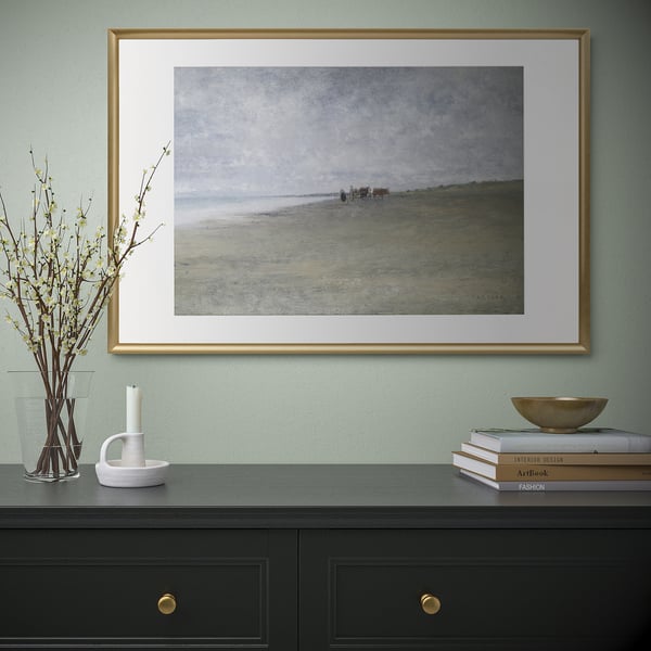 Gold-framed coastal picture on mint wall over black cabinet with gold handles, books, flowers, candle.