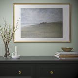 Gold-framed coastal picture on mint wall over black cabinet with gold handles, books, flowers, candle.