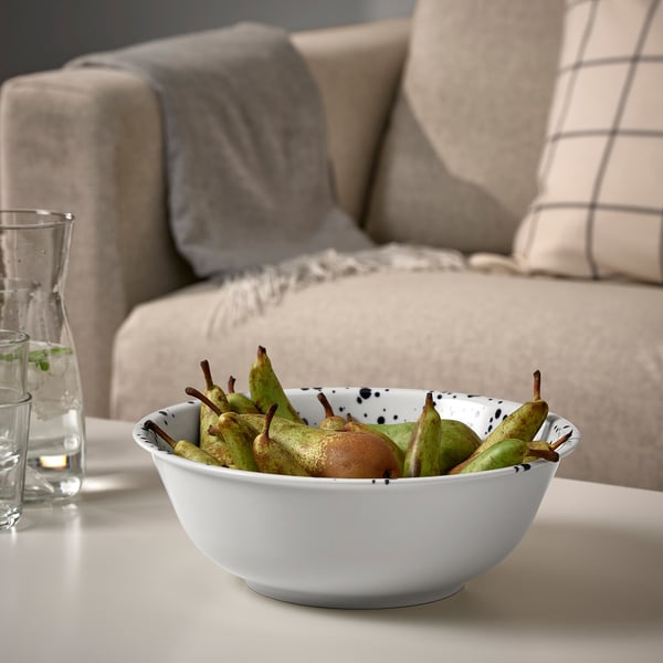 A bowl of green fruits sits on a table next to a glass pitcher. Behind it, a beige sofa with pillows.