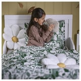 A girl in bed hugs a soft, roll-up hedgehog toy. Green patterned bedding and white flower pillows decorate the bed.