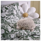 A grey SKOGSDUVA soft hedgehog toy on a green floral bed. It’s small, round, and plush, placed between large pillows.