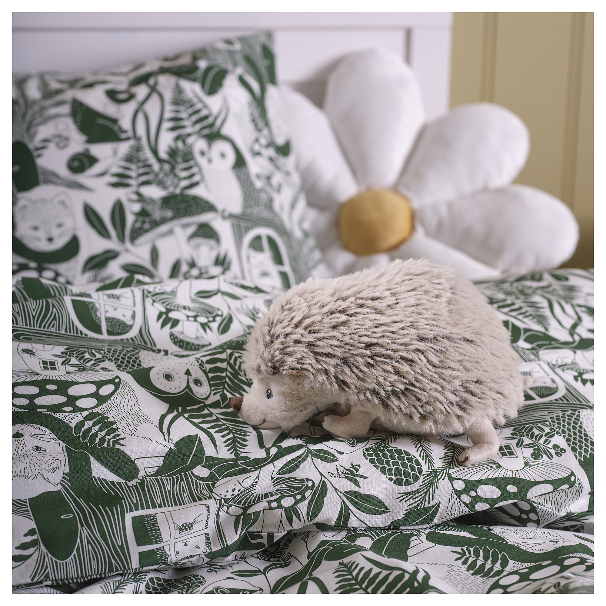 A grey SKOGSDUVA soft hedgehog toy on a green floral bed. It’s small, round, and plush, placed between large pillows.
