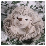 A plush toy hedgehog on patterned fabric, soft and detailed, with embroidered and a friendly, inviting smile.