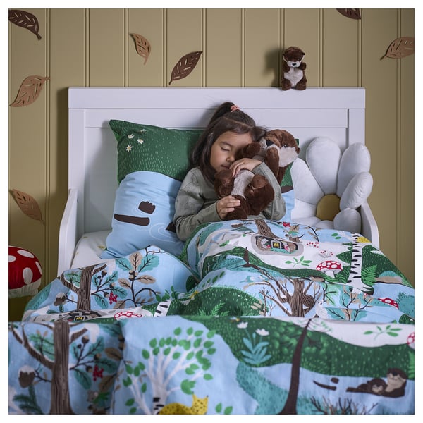 A child sleeps on a forest-themed bed, hugging two soft otter toys. The bedding and a wall decoration feature woodland motifs.