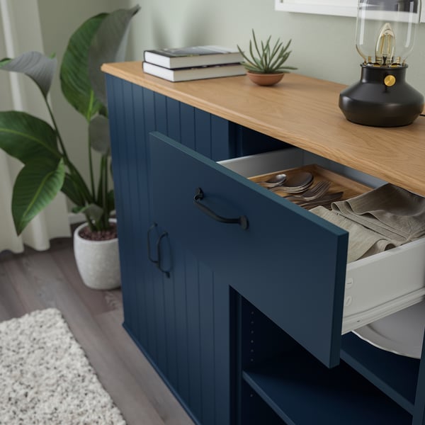 Blue sideboard with drawer open, displaying cutlery and napkins on wooden top beside potted plant.