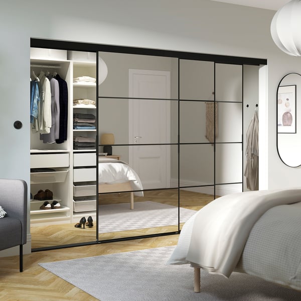 Modern bedroom with mirrored closet, bed, and chair reflected. Black frames, visible shelves, light wood floors, minimalist decor.