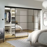 Modern bedroom with mirrored closet, bed, and chair reflected. Black frames, visible shelves, light wood floors, minimalist decor.