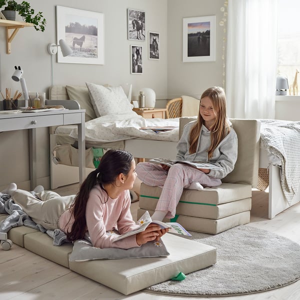 Two girls sit on modular floor cushions reading books in a cozy bedroom.