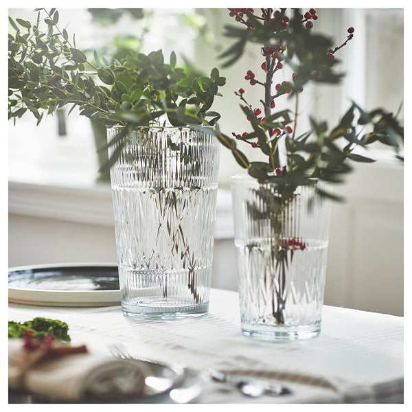 Two clear, vertically ridged vases hold greenery and red berries on a white tablecloth near a window, enhancing home decor with elegant simplicity.
