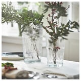 Two clear, vertically ridged vases hold greenery and red berries on a white tablecloth near a window, enhancing home decor with elegant simplicity.