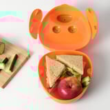 Bright orange SMASKA container with ears open, holding a sandwich and apple next to a wooden cutting board with cucumber slices.
