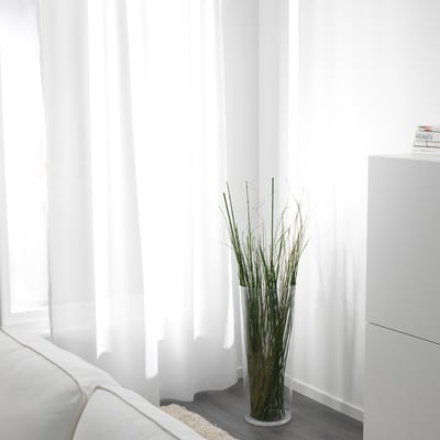 A minimalist living room with a white sofa; a vase of dried twigs stands near sheer white curtains, allowing natural light to filter through.