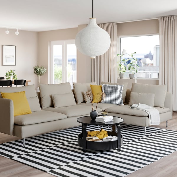 Modern living room with SÖDERHAMN sofa in beige, yellow & black pillows, striped rug, hanging lamp, large windows, plants, and coffee table.