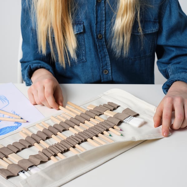 A person wearing a blue shirt uses a pencil case to organize art supplies. The case, made of light fabric, securely holds colored pencils and a brush.