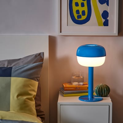 Bedside table with blue SOLHETTA lamp, books, and glass. Colorful pillows and art on wall.