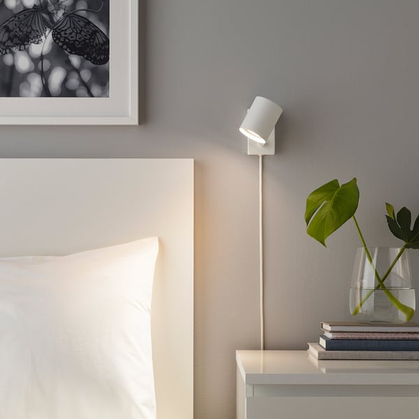 Minimalist bedroom with SOLHETTA lamp illuminating plant and books, gray walls, and butterfly print above bed.