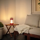 Red SOLVINDEN solar lantern on small wood table beside beige outdoor lounge chair.