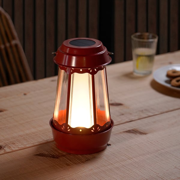 Red SOLVINDEN solar lantern on wooden table, emitting light, accompanied by a glass and cookies.