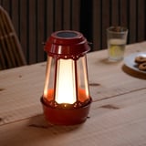 Red SOLVINDEN solar lantern on wooden table, emitting light, accompanied by a glass and cookies.