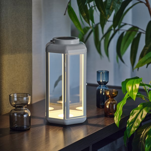 A modern, white solar-powered lantern sits on a wooden surface, emitting a warm glow. Its surrounded by small glass bottles and lush green plants.