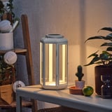Modern outdoor lamp on table, lit, with solar panel on top, surrounded by plants and decorations.