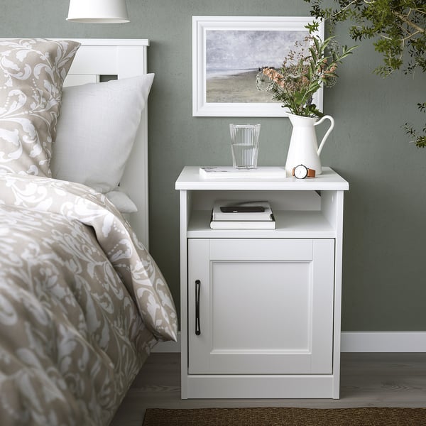 Small white SONGESAND bedside cabinet next to a bed. Holds books, glass, and flowers. Fits neatly beside the bed.