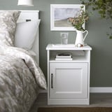 Small white SONGESAND bedside cabinet next to a bed. Holds books, glass, and flowers. Fits neatly beside the bed.