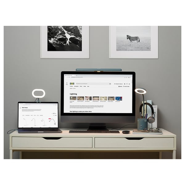 Modern home office with white desk, large monitor, laptop, ring lamp, headphones, and nature photos.