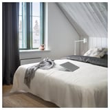 Modern bedroom with white bedspread, gray blanket, pillows, laptop, lamp, and large window showing trees.