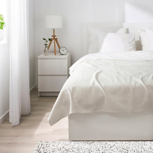 Bedroom scene: white STENFRÖMAL bedspread covers bed, wooden nightstand holds lamp, clock, and vase. Bright, minimalist decor with light wooden floor.