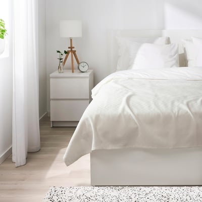 Bedroom scene: white STENFRÖMAL bedspread covers bed, wooden nightstand holds lamp, clock, and vase. Bright, minimalist decor with light wooden floor.