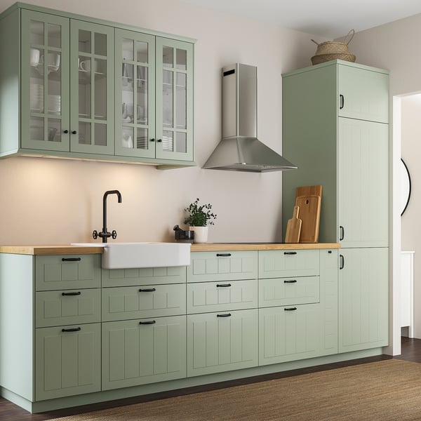 Modern kitchen with green cabinets, wood counter, white sink & black faucet. Tall units with glass doors and centered range hood.
