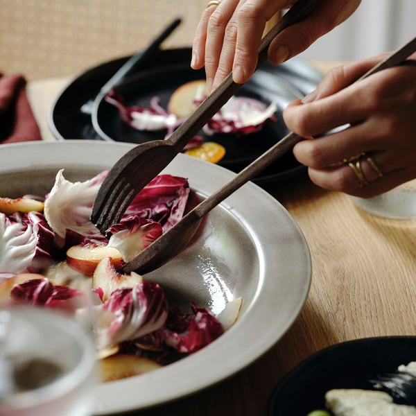Person holding wooden salad servers, placing reddish mixed salad onto a white plate, with pans in the background.