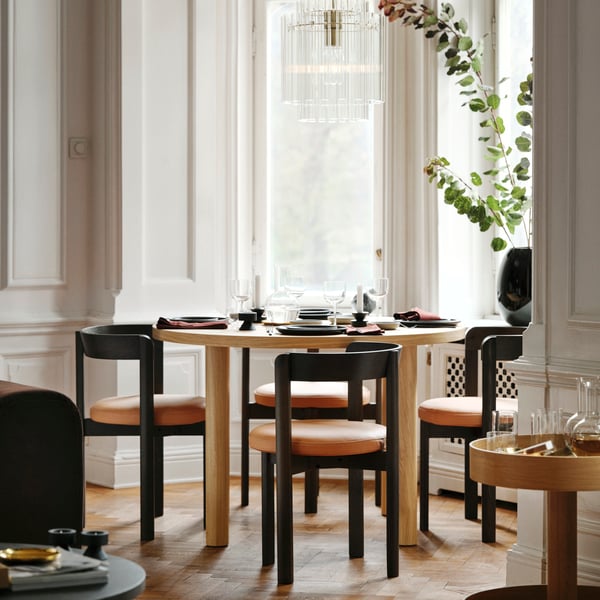 Oak dining set with black chairs, wine glasses, and eucalyptus vase by window.