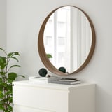 STOCKHOLM mirror: large round mirror with walnut frame, reflecting window. On small white dresser, its larger than books and plant. Decorative.
