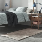 Bedroom with bed, nightstand, lamp, and beige rug. Neatly made bed with pillows. Nightstand has books and a lamp. Rug is soft and thick.