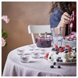 Table set with berry dessert, STÖRTSKÖN candles, glasses, stack of plates, and woman pouring drink.