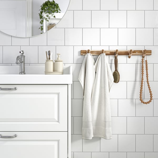 Modern bathroom with STOREDAMM wooden hook rack holding white robe. White tiles and cream dispensers. Acacia wood.