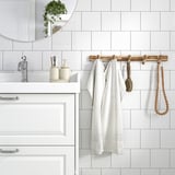 Modern bathroom with STOREDAMM wooden hook rack holding white robe. White tiles and cream dispensers. Acacia wood.
