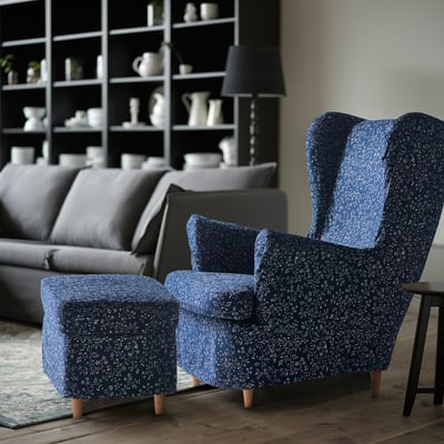 A cozy room with a blue-patterned STRANDMON wing chair and matching ottoman, positioned beside a gray sofa, with a bookshelf in the background.