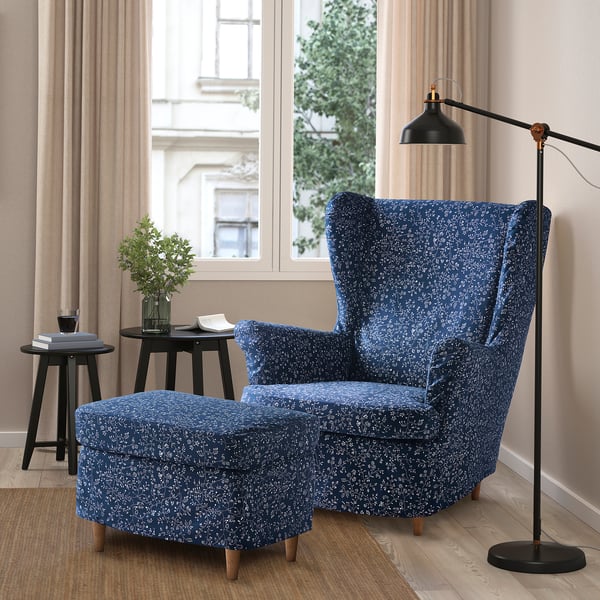 Modern living room with blue patterned chair, lamp, and side tables by large windows. Light flooring, beige curtains, city view.